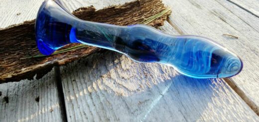 The Chrystalino Gallant dildo's glass is a cobalt blue that refracts the sunlight. It's resting on a white table against tree bark.