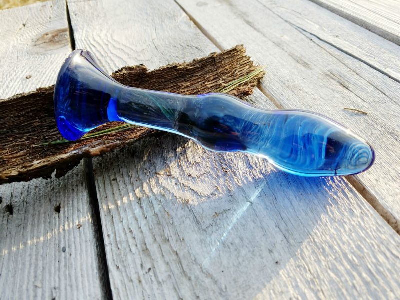 Top view. The Chrystalino Gallant dildo's glass is a cobalt blue that refracts the sunlight. It's resting on a white table against tree bark.