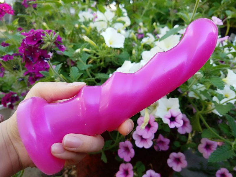 Holding a strawberry pink Tantus Tsunami in my hand amid purple, white, and pink flowers. Ridges' side view.