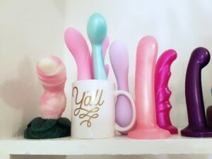 [Image: BMS Pillow Talk Sassy in a mug with other vibes and next to silicone dildos in my closet]