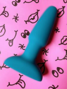 B-Vibe Rimming Plug on pink doodle background with tongues, lips, and buttholes