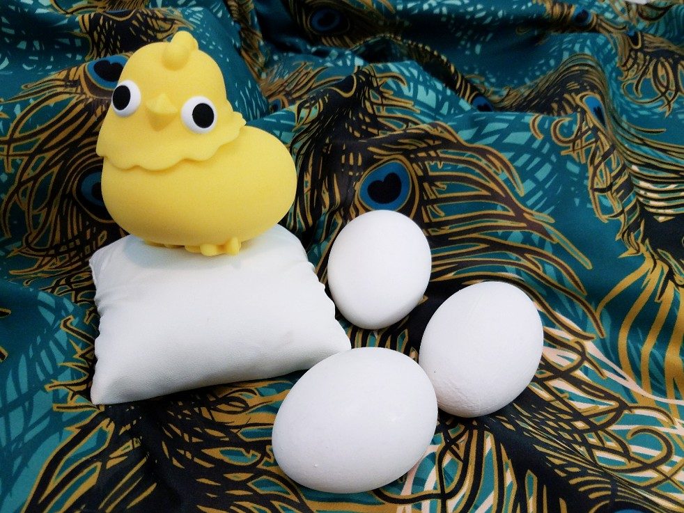 Image: Emojibator Chickie clitoral suction vibrator resting on a pillow next to large eggs. Its body is about the same size as an egg.