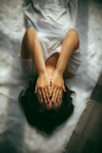 [Image: a woman in a white V-neck lying in bed, covering her face with her hands]