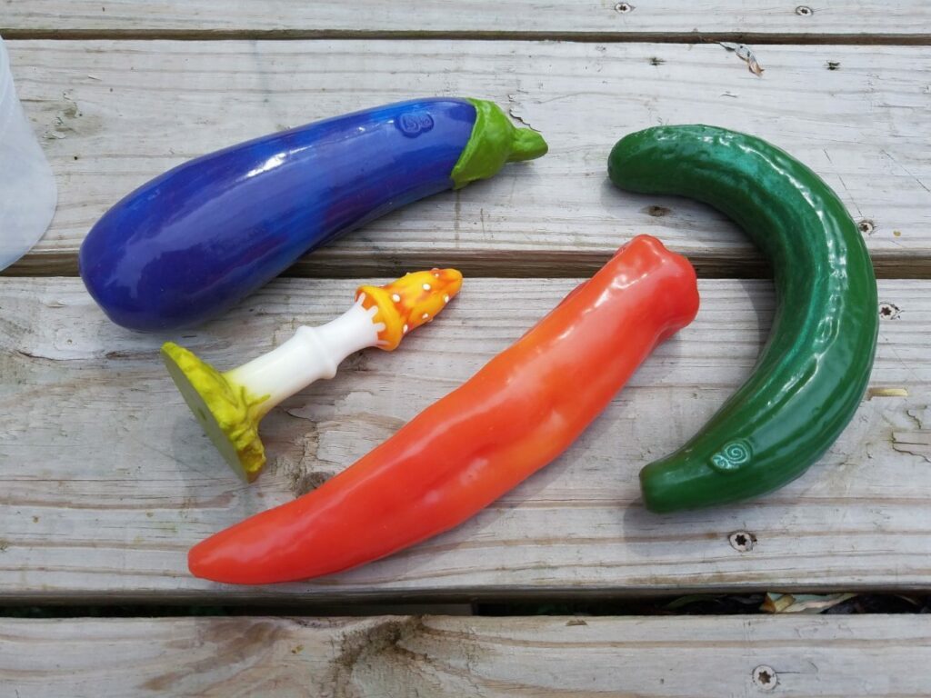 [Image: girthy and smooth Self Delve Eggplant / Aubergine next to Fly Agaric butt plug next to red pepper dildo next to curved cucumber dildo. They're all thermochromic / color-changing pigments in glossy silicone!]