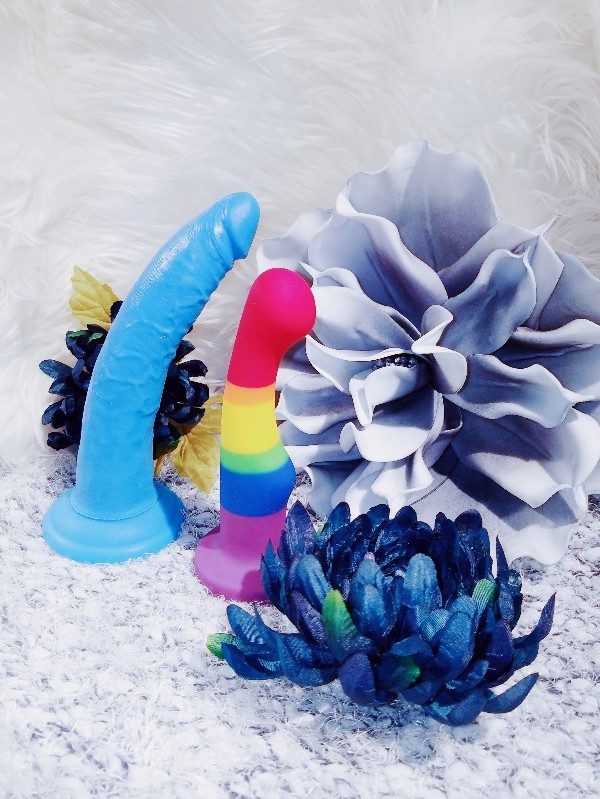 [Image: blue Blush Neo Elite 7.5" dual-density silicone dildo with suction cup base and without balls next to Blush Avant Pride P1 rainbow striped dildo in front of white faux fur background and next to blue and white flowers]
