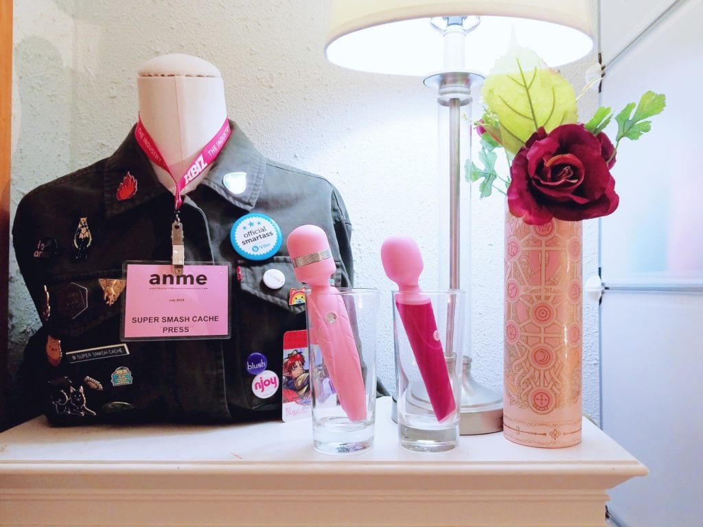 [Image: Pink BMS Factory Pillow Talk Cheeky and Blush Noje W4 Wand vibrators on display in cups in front of a mannequin with jacket and enamel pins]