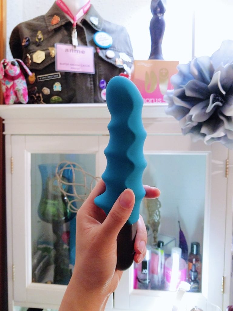 [Image: Petrol Blue/Teal Fun Factory Stronic Surf Pulsator in my hand, in front of my condom and lube cabinet. The lighting shows much contrast along the shaft's waves]