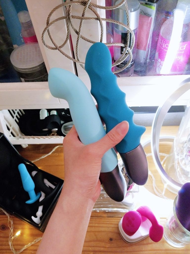 [Image: holding the Pool Blue Fun Factory Stronic G and Petrol Stronic Surf pulsators side view in front of my lube and condom cabinet with some of the other toys on the shelf visible]