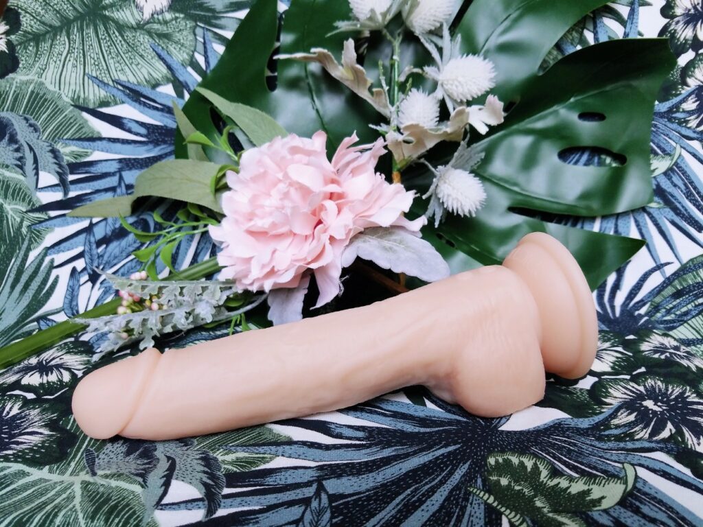 [Image: BMS Factory Naked Addiction realistic dual-density self-thrusting dildo with suction cup base next to leaves and flowers horizontal view]