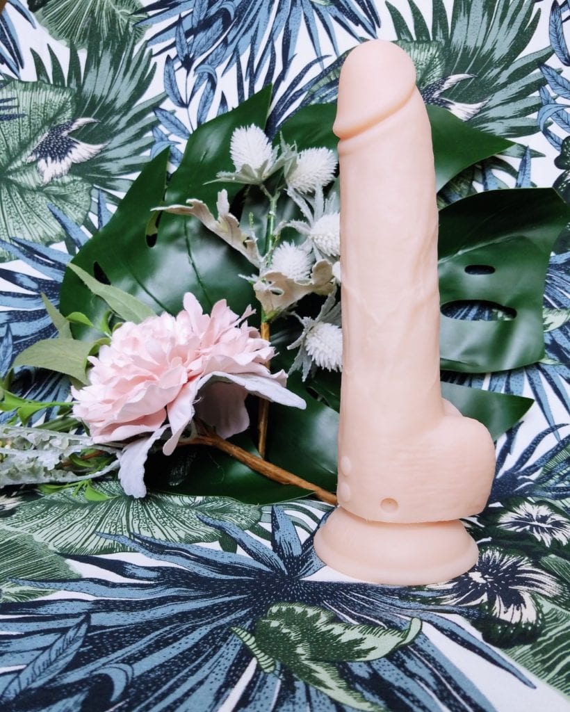 [Image: BMS Factory Naked Addiction realistic dual-density self-thrusting dildo with suction cup base next to leaves and flowers vertical view]