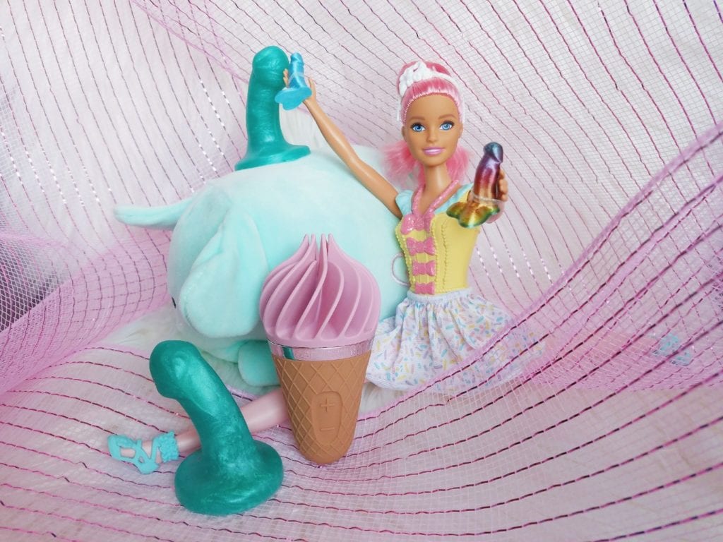 [Image: Barbie doll doing a split among Uberrime silicone teenie weenies, holding Barbie-sized dildos next to pink and brown Satisfyer Sweet Treat rotating ice cream clitoral stimulator]