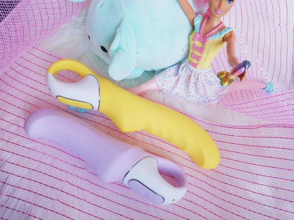 [Image: purple Satisfyer Charming Smile G-spot vibrator and Yummy Sunshine yellow textured vibrator next to Barbie doll doing a split and holding a tiny rainbow dildo]
