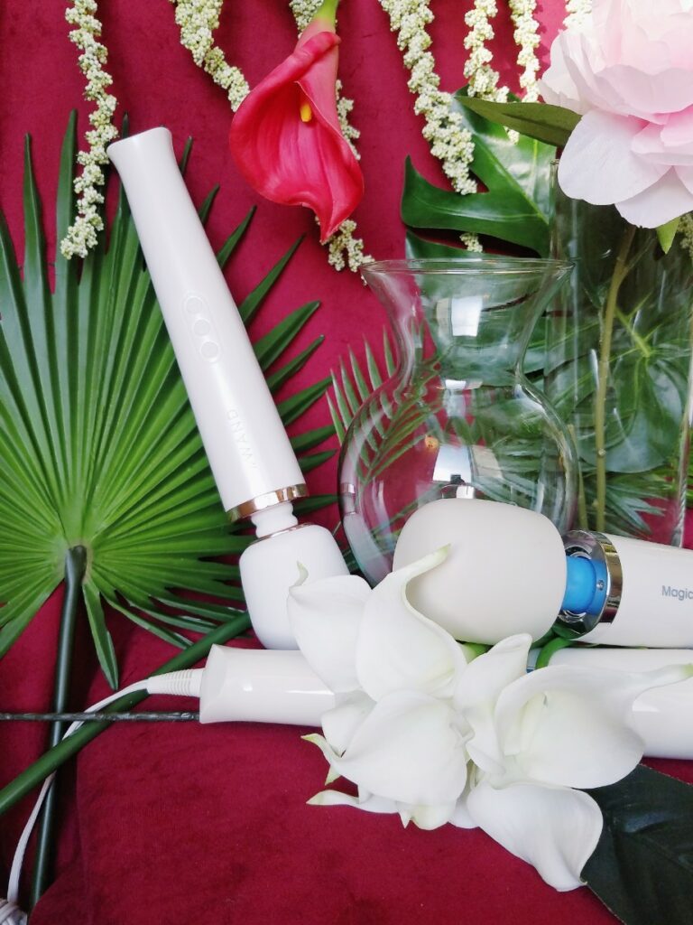 [Image: Le Wand Rechargeable next to vase, Magic Wand Plus, and glass vase among plants and tropical flowers]