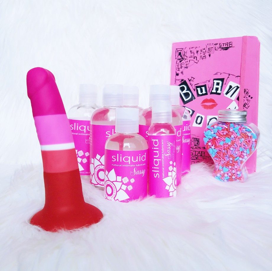 [Image: Blush Novelties Avant Pride P3 Beauty/Lesbian Pride striped dildo with MANY bottles of different sizes of Sliquid Sassy with pink label, plus Mean Girls Burn Book journal and bottle of candy sprinkles for decoration]