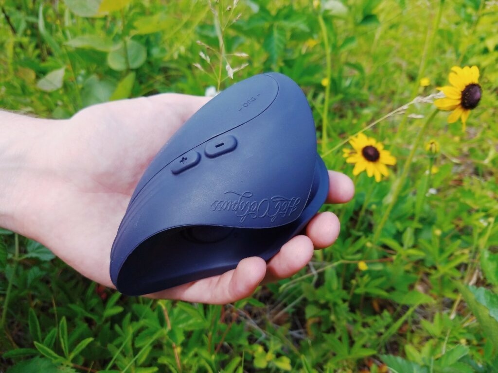 [Image: hand holding Hot Octopuss Pulse Duo penis vibrator in a grassy background with yellow flowers]