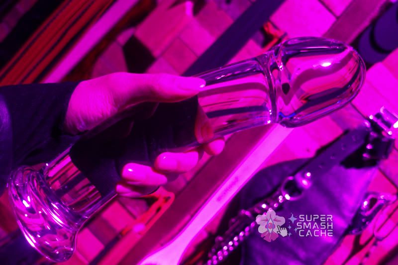 Extra large realistic mushroom tip glass dildo in hand in front of impact toys on wall